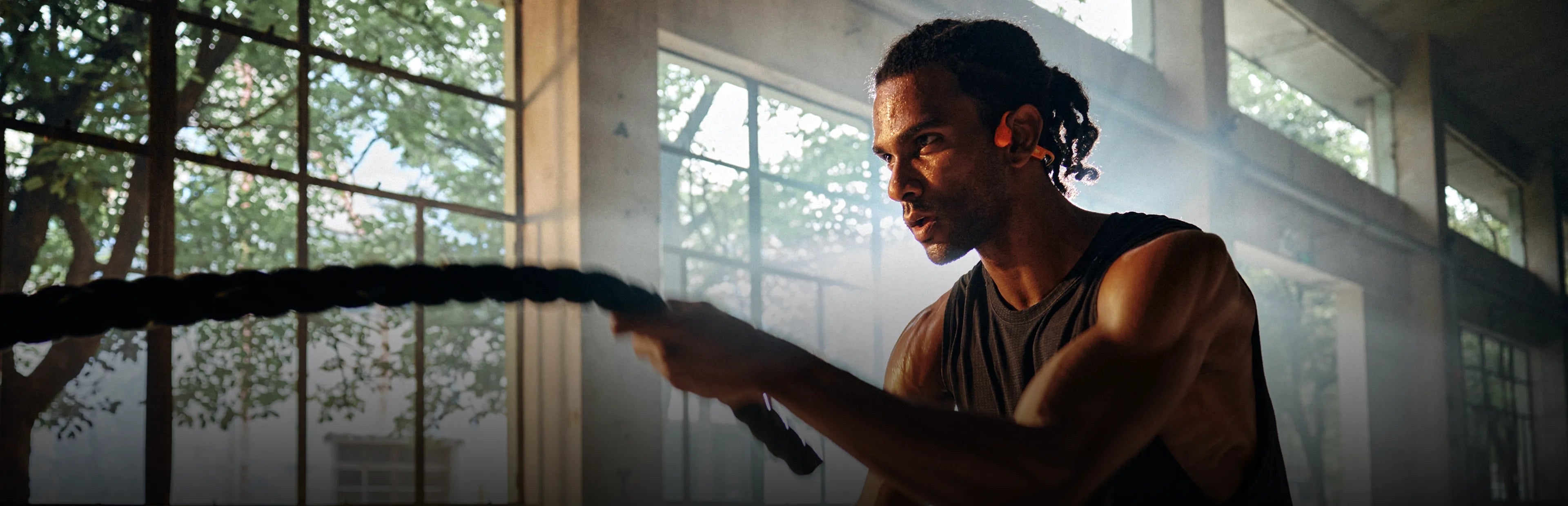 Open-Ear Headphones.Made for Every Sport. Man holding a rope in front of large windows with natural light.