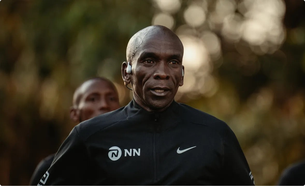 Eliud Kipchoge World Record Holder in Marathon wearing a black jacket with NN and Nike logos outdoors. Promote Bone Conductible headphones