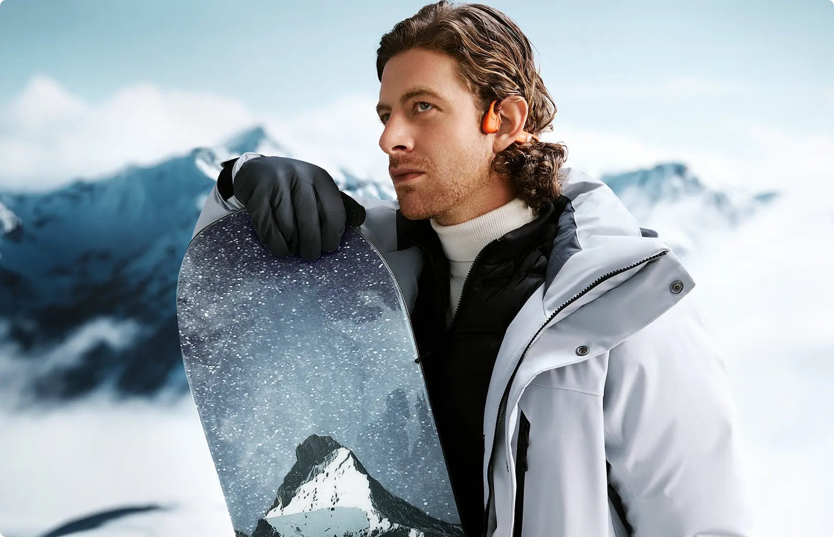 Man wear ear open Bone Conductible Headphones holding a snowboard with mountains in the background